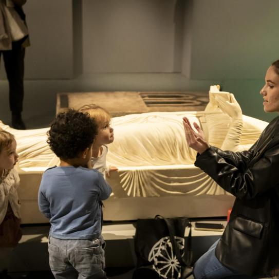 Photographie d'enfants autour d'une femme mimant le gisant d'Aliénor d'Aquitaine qui tient dans ses mains un livre ouvert